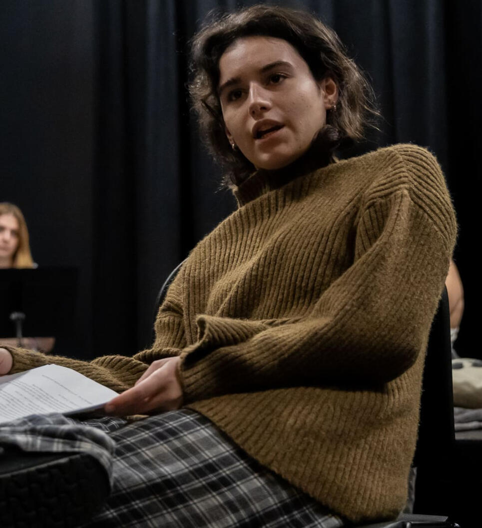 Photo of a young woman with dark hair and a yellow sweater performing a play.