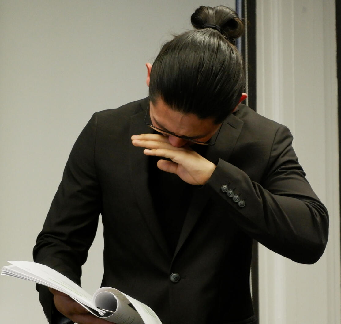 Photo of a man with black hair holding a script, looking stressed or pained.